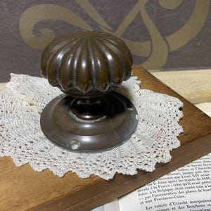 May include: A vintage bronze door knob with a fluted, rounded top and a circular base. It rests on a white lace doily, placed on a wooden surface. The knob has a dark, aged patina.