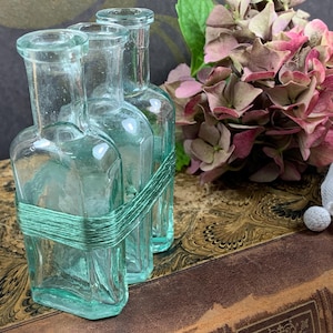 Op de afbeelding: Drie heldere glazen flessen met een lichtblauwe tint zijn gegroepeerd, gebonden met turquoise touw. Een boeket roze en mauve hortensia's en een wit kanten kleedje staan op de achtergrond, wat een vintage esthetiek creëert.