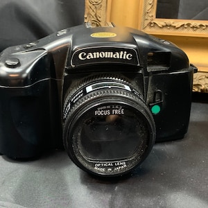 May include: A black Canonmatic 35mm film camera with a 50mm 1.6.3 focus free lens. The lens has the text "Optical Lens Made in Japan" printed on it.