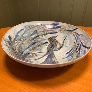 Könnte beinhalten: Eine große, ovale Servierschüssel aus Keramik mit handgemaltem Vogel- und Blattdesign. Die Schüssel hat einen hellblauen Hintergrund mit blauen, grünen und braunen Akzenten. Der Vogel sitzt auf einem Ast.