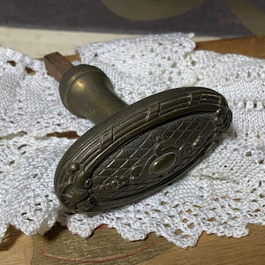 May include: An antique bronze door handle with an oval, ornate design. The handle features detailed patterns and a dark patina, resting on a white lace doily. The handle's design is reminiscent of vintage hardware.