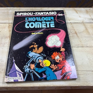 May include: A vintage comic book cover featuring the characters Spirou and Fantasio. The cover is black with a pink comet and a white object in the centre. The title of the comic is "L'Horloger de la Comete" and the text "Tome 8: Janry" is below the title. The comic is published by Dupuis.