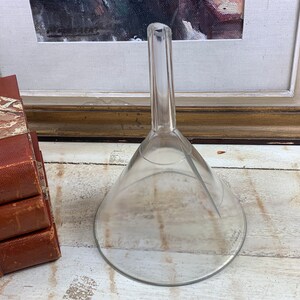 May include: A clear glass funnel with a long, narrow spout. The funnel is sitting on a white wooden surface.
