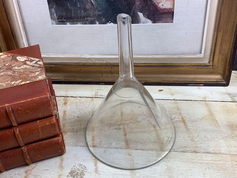 May include: A clear glass funnel with a long, narrow spout. The funnel is sitting on a white wooden surface.