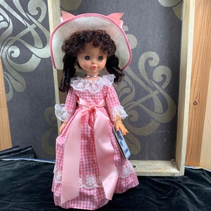 Puede incluir: Un vestido de cuadros vichy rosa y blanco con una cinta de raso rosa y ribete de encaje. La mu&ntilde;eca lleva un sombrero blanco con un lazo rosa y tiene el pelo rizado marr&oacute;n.