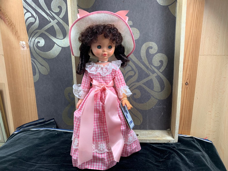 Puede incluir: Un vestido de cuadros vichy rosa y blanco con una cinta de raso rosa y ribete de encaje. La mu&ntilde;eca lleva un sombrero blanco con un lazo rosa y tiene el pelo rizado marr&oacute;n.