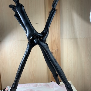 Op de afbeelding: Een zwart houten beeld van twee in elkaar verweven figuren, waarvan de armen en benen een driepotige basis vormen. De figuren hebben gestileerde menselijke kenmerken en zijn met ingewikkelde details gesneden.