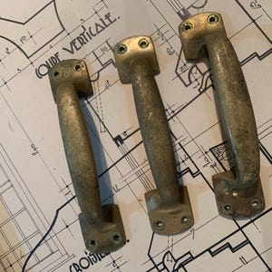 Könnte beinhalten: Drei antike Türgriffe aus Messing mit verwittertem Finish, angeordnet auf einem alten Architekturplan. Die Griffe haben ein gebogenes Design mit Montageplatten an jedem Ende. Der Bauplan trägt die Aufschrift "GUIDE VERTICALE."