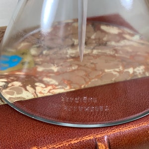 May include: A clear glass funnel with a round base and a spout. The funnel has the text "Berggione" and "Art Vindel" printed on the bottom.
