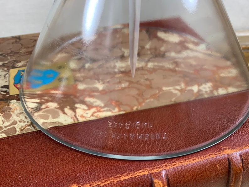 May include: A clear glass funnel with a round base and a spout. The funnel has the text "Berggione" and "Art Vindel" printed on the bottom.