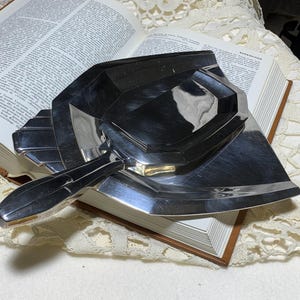 May include: A vintage, Art Deco-style silver-toned ashtray with a geometric design, resting on an open book. The ashtray features a handle and a series of stepped, angular elements. The book has aged pages and a brown cover.