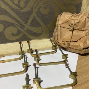 May include: A collection of brass drawer pulls with mounting hardware, arranged on a white surface. A bundle wrapped in brown paper is in the background. The pulls have a curved handle design and are likely vintage or antique.