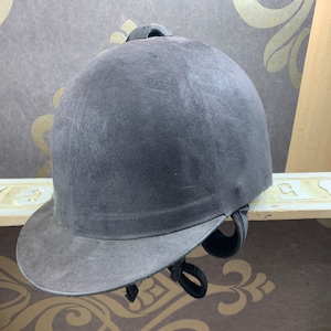 May include: A black riding helmet with a suede finish and a black brim. The helmet has a black adjustable strap.