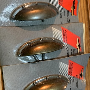 May include: Three antique bronze cabinet knobs with a rounded, oval shape and a decorative row of small studs around the edge. The knobs are displayed on packaging with the text "Collezione Ingresso 615692 1 stp" in red and black.