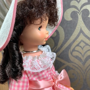 Puede incluir: Una mu&ntilde;eca vintage con cabello casta&ntilde;o rizado, vestida con un vestido de cuadros vichy rosa y blanco con un cuello de encaje blanco, una cinta de raso rosa y un sombrero blanco con ribete rosa.