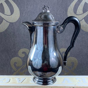 Op de afbeelding: Een zilveren koffiekan met een zwarte handgreep en een decoratief deksel. De kan staat op een wit oppervlak met het woord "BEV" erop gedrukt.