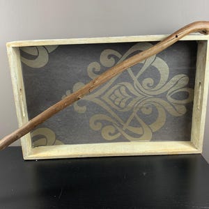 May include: A wooden walking stick with a curved handle rests on a decorative tray. The tray has a cream-colored frame and a dark gray interior with a gold floral pattern. A small wooden container sits to the left.