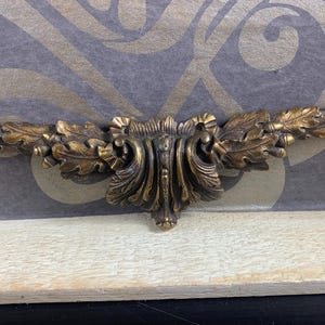 Könnte beinhalten: Antike, bronzefarbene Zierleiste mit aufwendigem Design. Die Leiste zeigt ein zentrales, stilisiertes Blumendesign, flankiert von detaillierten Blatt- und Eichelmotiven. Der Hintergrund ist grau und gold gemustert.