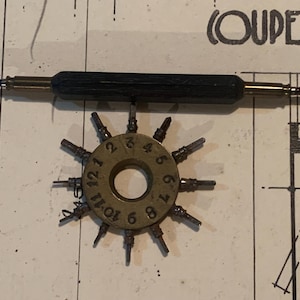 May include: A vintage watch repair tool set. The set includes a double-ended screwdriver with a black handle and a circular tool with numbered posts. The tools are made of metal and are set against a blueprint background.