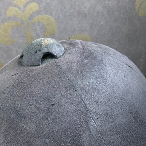May include: A gray riding helmet with a black leather strap and a black buckle. The helmet has a textured surface and a small vent on the top.