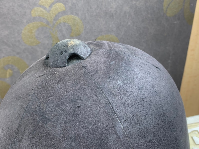 May include: A gray riding helmet with a black leather strap and a black buckle. The helmet has a textured surface and a small vent on the top.
