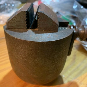 May include: A small, gray metal vise with a black rubber jaw pad. The vise is mounted on a round base.