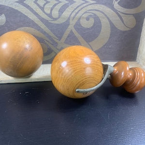 May include: Three wooden drawer pulls. Two are spherical, one is a complete pull with a metal handle and two wooden knobs. The wood has a light brown colour and visible grain. The pulls are on a black surface.