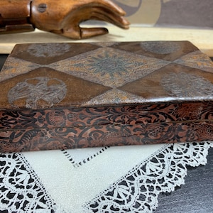 Peut inclure: Boîte rectangulaire en cuir marron antique avec des motifs floraux et géométriques en relief. Le couvercle de la boîte présente un motif en losange. Posée sur un napperon en dentelle blanche.