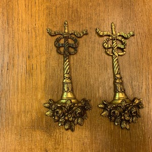 Pode incluir: Um par de ornamentos de parede em metal dourado a condizer, com um design de coluna vertical canelada rematada por um laço decorativo e terminada na base com um relevo detalhado de rosas e folhas agrupadas.