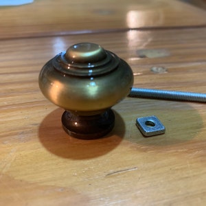 Peut inclure: Un bouton de porte de couleur or avec un dessus rond et bombé et une base carrée. Le bouton est livré avec une vis et une rondelle carrée.