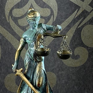 May include: A bronze statue of Lady Justice, blindfolded, holding scales and a sword. The statue has a verdigris patina with gold accents. The background is a dark gray with a repeating gold pattern.