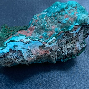 Puede incluir: Una roca rugosa de forma irregular con un depósito mineral azul verdoso, verde y marrón. El depósito mineral tiene un aspecto estratificado y estriado.