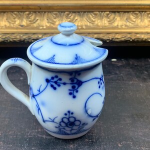 Op de afbeelding: Een kleine witte porseleinen roomkan met een blauw-wit bloemenmotief. De roomkan heeft een deksel met een klein tuitje.