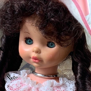 Puede incluir: Una mu&ntilde;eca de porcelana con el pelo largo y rizado de color marr&oacute;n oscuro, que lleva un sombrero blanco con ribete rosa y un vestido de encaje rosa y blanco. La mu&ntilde;eca tiene ojos azules y un collar de plata.