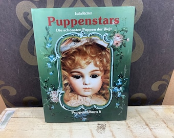 May include: A green book with the title "Puppenstars" and the subtitle "Die schönsten Puppen der Welt". The cover features a portrait of a porcelain doll with blonde hair and blue eyes. The book is part of a series, as indicated by the text "Puppen Album 6" at the bottom.