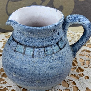 Op de afbeelding: Een blauwe en witte keramische kan met een getextureerd oppervlak en een geometrisch ontwerp rond het midden. De kan heeft een handvat en een rond lichaam.