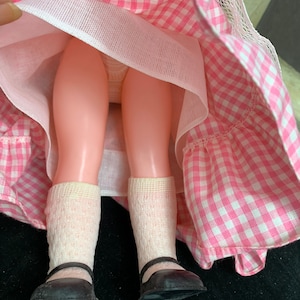Puede incluir: Una mu&ntilde;eca vintage que lleva un vestido de cuadros vichy rosa y blanco con un enaguas blanco y zapatos negros.