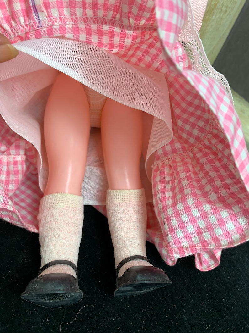 Puede incluir: Una mu&ntilde;eca vintage que lleva un vestido de cuadros vichy rosa y blanco con un enaguas blanco y zapatos negros.