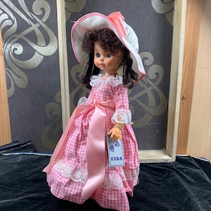 Puede incluir: Un vestido de cuadros vichy rosa y blanco con ribete de encaje blanco y una cinta de raso rosa. La mu&ntilde;eca lleva un sombrero blanco con una cinta rosa y tiene el pelo rizado marr&oacute;n. La mu&ntilde;eca sostiene una etiqueta que dice "FIBA".