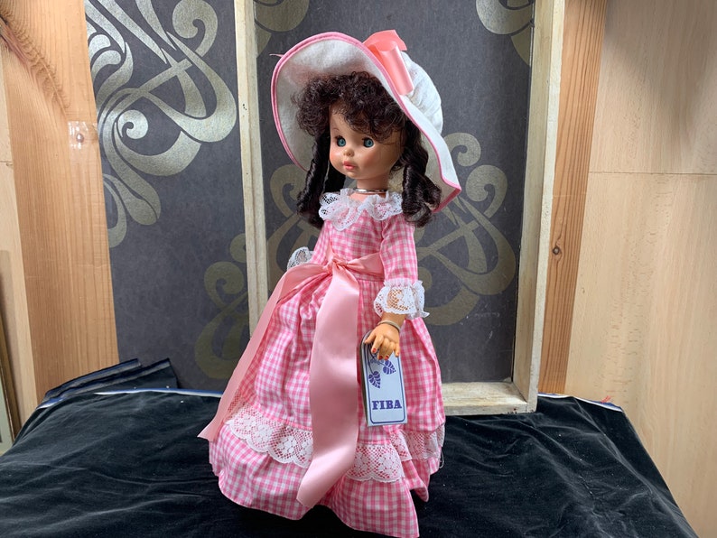 Puede incluir: Un vestido de cuadros vichy rosa y blanco con ribete de encaje blanco y una cinta de raso rosa. La mu&ntilde;eca lleva un sombrero blanco con una cinta rosa y tiene el pelo rizado marr&oacute;n. La mu&ntilde;eca sostiene una etiqueta que dice "FIBA".