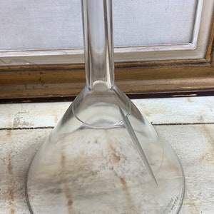 May include: A clear glass funnel with a wide, flared opening and a long, narrow stem. The funnel is sitting on a light-colored wooden surface.