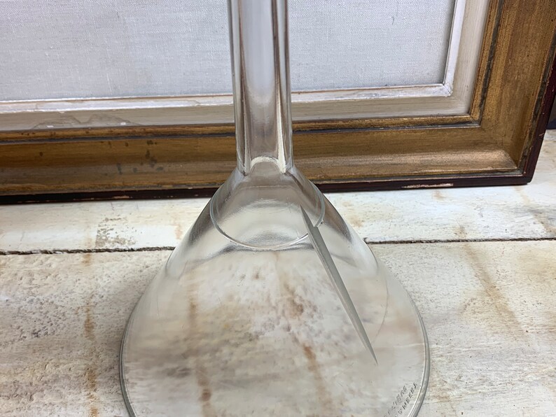 May include: A clear glass funnel with a wide, flared opening and a long, narrow stem. The funnel is sitting on a light-colored wooden surface.