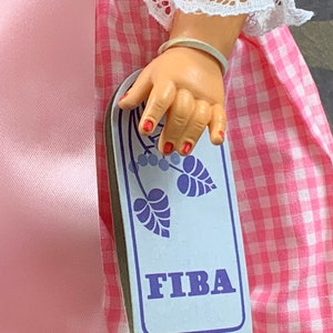 Puede incluir: Primer plano de la mano de una mu&ntilde;eca sosteniendo una tarjeta rectangular blanca con un dise&ntilde;o floral azul y la palabra "FIBA" impresa en letras azules.