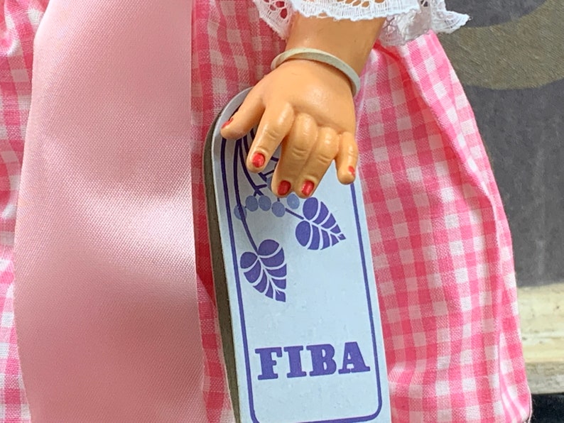 Puede incluir: Primer plano de la mano de una mu&ntilde;eca sosteniendo una tarjeta rectangular blanca con un dise&ntilde;o floral azul y la palabra "FIBA" impresa en letras azules.