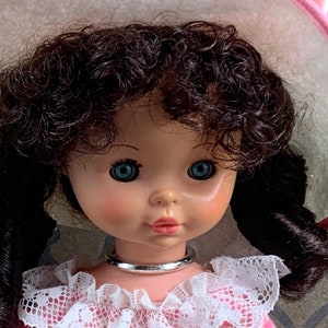 Puede incluir: Una mu&ntilde;eca de porcelana con cabello casta&ntilde;o oscuro rizado, ojos azules y un sombrero rosa y blanco. La mu&ntilde;eca lleva un vestido rosa con encaje blanco y un collar plateado.