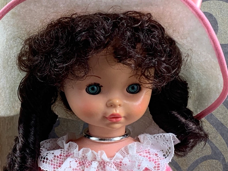 Puede incluir: Una mu&ntilde;eca de porcelana con cabello casta&ntilde;o oscuro rizado, ojos azules y un sombrero rosa y blanco. La mu&ntilde;eca lleva un vestido rosa con encaje blanco y un collar plateado.