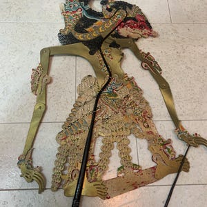 Op de afbeelding: Een traditionele Indonesische Wayang Kulit-pop. De pop is gemaakt van goudkleurig leer met ingewikkelde uitgesneden ontwerpen en geschilderde details in rood, blauw en groen. Het heeft beweegbare ledematen en wordt vastgehouden met zwarte staven. De pop ligt op een lichtgekleurd oppervlak.