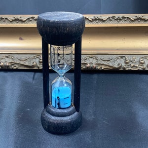 Puede incluir: Un reloj de arena de madera negra con arena azul. El reloj de arena tiene un bulbo de vidrio con marcas para medir el tiempo.