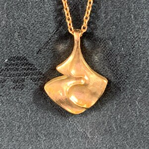 Puede incluir: Un collar con un colgante de tono dorado con un diseño abstracto único. El colgante tiene la forma de una flor o una hoja estilizada, con una textura lisa y orgánica.