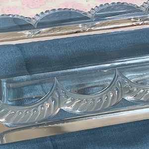 Puede incluir: Una bandeja rectangular de vidrio transparente con borde festoneado. La bandeja tiene un diseño curvo decorativo en los lados. El vidrio transparente permite el paso de la luz. Probablemente se utiliza con fines decorativos.